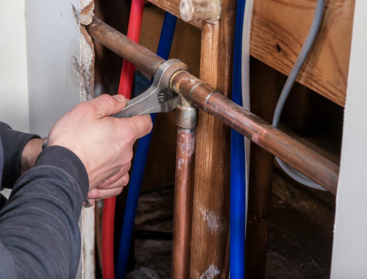 Plumber tightening a copper pipe fitting beside PEX water lines inside a wall
